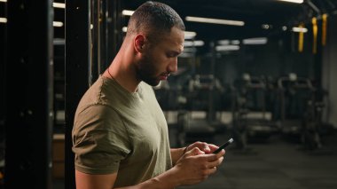 Afrika kökenli Amerikalı sporcu cep telefonlu erkek fitness eğitmeni spor salonunda spor yaptıktan sonra rahatlıyor akıllı telefon teknolojisini tarıyor internet mesajlarına bakıyor kameraya bakıyor