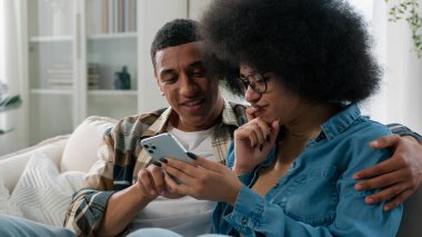 Mutlu evli Afro-Amerikan çift kanepe görünümlü adam ve kadın cep telefonu alışveriş web sitesi uygulaması internet Akıllı telefon arama biletleri kullanarak konuşma tartışarak satın alma