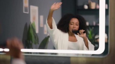 Aynaya bakan neşeli Afro-Amerikan kadın saç fırçasında şarkı söylüyor mikrofon gibi karaoke söylüyor banyoda dans ediyor banyoda eğleniyor banyoda kız kız arkadaşının evinde eğleniyor
