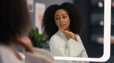 Gülümseyen güzel Afro-Amerikan mutlu sağlıklı kadın evde pohpohlanıyor cilt teması saç dokunuşu kozmetik güzellik prosedürü aynadaki yansıma kadın maske kremi kozmetik kıza banyo yaparken şımartıyor.