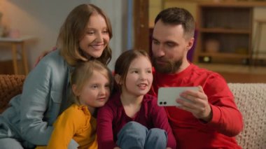 Mutlu bir aile, Kafkasyalı ebeveynler, cep telefonlarıyla video konuşmaları, neşeli çocuklar, akıllı telefonlar, anne babalar, küçük kızlar, kız kardeşler, ev rekorları blogunda selfie çekiyorlar.