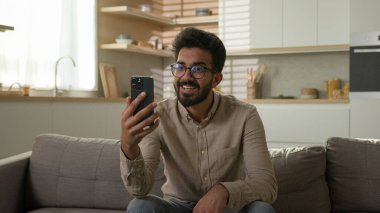 Gülümseyen Hintli, Arap, etnik erkek, serbest çalışan, mutlu blogcu, ev sahibi, konuşma, cep telefonu görüşmeleri, akıllı telefon görüşmeleri, online blog sohbetleri, mutfak kanepesinde.