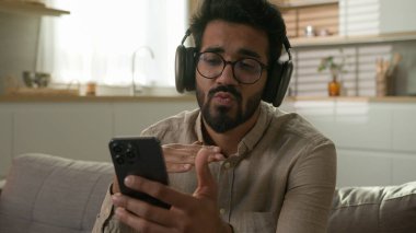 Kaygısız, rahat Arap erkek, kulaklıklı, etnik erkek, müzik dinleyen kulaklıklı, kablosuz kulaklıklı, disko müzikli, ev mutfağındaki cep telefonlu, çevrimiçi ses servisinin tadını çıkarıyor.