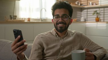 Arap Hintli erkek, etnik ev sahibi, neşeli, mutlu adam, ev mutfağında cep telefonuyla video film izliyor, haberleri okuyor, sabah kahvesini içiyor, kanepede dinleniyor, kamera tebessümüne bakıyor.