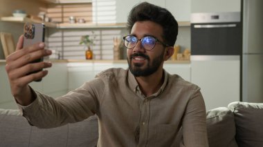 Gülümseyen Hintli, Arap, etnik erkek, serbest çalışan, mutlu blogcu, ev sahibi, konuşma, cep telefonu görüşmeleri, akıllı telefon görüşmeleri, online blog sohbetleri, mutfak kanepesinde.