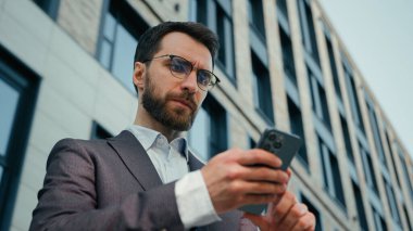 Ciddi bir Kafkasyalı girişimci yetişkin erkek işadamı CEO iş adamı ofis yöneticisi açık havada akıllı telefona bakarak şehirdeki sosyal ağları tarayıp cep telefonuyla sohbet ediyor.