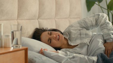 Yatak odasında yatan bir kadın karnını tutarak acı çekiyor. Karın ağrısı, mide ağrısı, sindirim bozukluğu, regl, sağlıksız regl ağrıları, karın ağrısı.