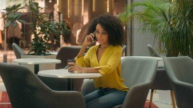 Genç Afrikalı Amerikalı kadın kafede oturup kahve içiyor cep telefonuyla konuşuyor cep telefonlarıyla sohbet ediyor alışveriş merkezini arıyor hafta sonu dinleniyor gülümseyen kadın iletişimi kuruyor zarifçe konuşuyor.