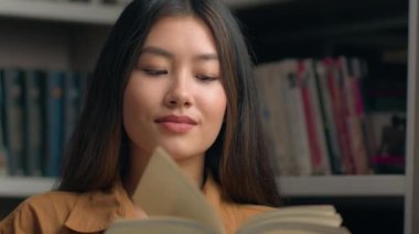 Üniversite kütüphanesinde kitaplığın yanında kitap okuyan Asyalı kadın portre okuyucusu. Akıllı odaklı Koreli kız öğrenci. Okuma kitabı okuyor.