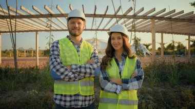 Takım çalışanı üniforması Kafkasyalı mühendis kadın mimar kadın kız müteahhit erkek ustabaşı kameraların çapraz kollarına bakıyor açık hava parkında el ele poz veriyor.