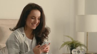 Kafkasyalı bir kadın elinde cep telefonu akıllı telefon mesajları mesajlaşarak mutlu, mutlu, mutlu, gülümseyen ev odası teknolojisi ile internet iletişiminin keyfini çıkarıyor.