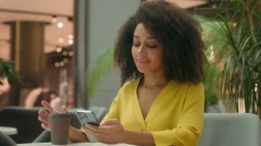 Sıcak kahve molası veren mutlu Afro-Amerikalı kadın rahatla mesajlaşarak akıllı telefon görüşmeleri yap cep telefonu gülümse hobi yap alışveriş merkezi kafeteryasında sakin bir bayan