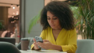 Ciddi Afrika kökenli Amerikalı kadın cep telefonu akıllı telefonu kullanıyor. Kız, ekran ekranına bakıyor. Kahve içiyor, karmaşıklaşıyor.