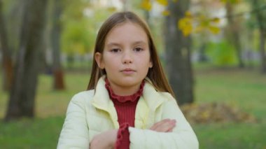 Kafkasyalı küçük kız parkta el ele tutuşup kollarını göğsünde kavuşturdu. Kendine güveni tam, üzgün kız öğrenci kız öğrenci. Sonbahar parkı mevsiminde çocuk bakımı.