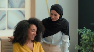 İki mutlu iş kadını kız çok ırklı iş kadınları ofiste çalışıyor Arap Müslüman kadın lider tesettüre Afrikalı Amerikalı öğrencilere yardımcı oluyor gülümsüyor