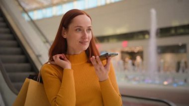 Genç kaygısız Kafkasyalı kadın kadın müşteri müşteri müşterisi iş kadını kağıt çantalar alıyor cep telefonundan sesli mesajlar alıyor kapalı kapılar ardında alışveriş merkezindeki alışveriş merkezlerinde sesli mesajlar gönderiyor.