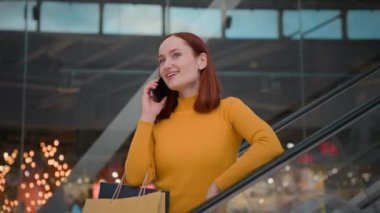 Kahkahalar atan Kafkasyalı, kızıl saçlı, iş kadını, yürüyen merdivene binen kadın, elinde hediye çantalarıyla mutlu bir kadının içinde akıllı telefon, cep telefonu, güler yüzlü alışveriş merkezi.