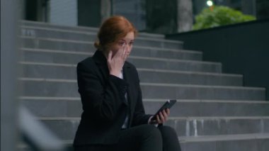 Hüsrana uğramış Kafkasyalı genç iş kadını, kızıl, kadın, elinde cep telefonu, akıllı telefon, negatif mesaj, olumsuz sohbet, iş kaybı, iflas, işsizlik merdivenlerinde belediye binasının önünde oturuyor.