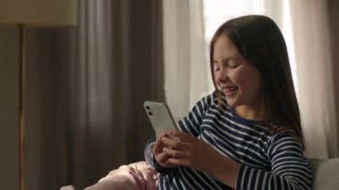 Kafkasyalı mutlu küçük kız koltukta uzanmış cep telefonu akıllı telefon zımbırtısı çocuk çocuk çocuk teknolojisi mesajlaşması internet zevki gülümsemesi internet siteleri kadın iletişimi