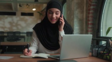 Mutlu Müslüman İslamcı kadın tesettürlü kadın Arap kadın çoklu görev sahibi iş kadını yöneticisi konuşuyor cep telefonu cevapları akıllı telefon görüşmeleri danışma istemcisi ofisteki bilgisayarda yazı yazan notlar yazıyor.