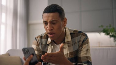 Öfkeli Afrikalı Amerikalı erkek melez erkek, etnik erkek kullanıcı sinirli, sinirli, bağıran, bağıran, bağıran, cep telefonlu, görüntülü, sohbet konferansı tartışması ev kanepede tartışmalı.