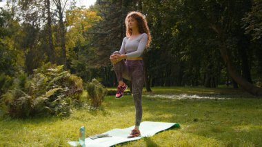 Caucasian athlete sportswoman woman standing on one leg balancing holding pressing knee to chest sport stretching yoga balance stretch flexibility workout yoga on mat in park at nature girl exercise clipart