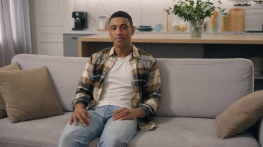 Eve portre mutlu ruh hali olumlu etnik adam gülümsüyor rahat kanepe dinlenme hafta sonu kameraya bakıyor sağlıklı Afrikalı Amerikalı erkek erkek erkek erkek gülümseme kanepede dişlek gevşek kiracı