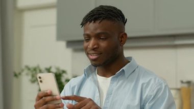 Mutlu gülümseyen Afro-Amerikan etnik erkek kullanıcı şampiyonu çift ırklı erkek etnik erkek gülümsemesi kazandı akıllı telefon kazandı zafer elde edildi iyi haberler ev mutfağında cep telefonu neşesi