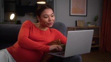 Mutlu gülümseyen Afro-Amerikan hamile kadın hamile anne hamileliği ev kanepede keyfine varıyor dizüstü bilgisayarla sohbet ederek internetten bilgisayar alışverişi yaparak.