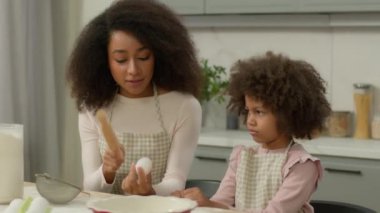 Afro-Amerikan aile şefkatli anne anne anne anne küçük kız çocuğa yemek pişirmeyi öğretiyor un buğday buğdayını gösteriyor yumurta gösteriyor merdaneyi veriyor mutfakta konuşan aşçıyı nasıl kullanacağını açıkla.