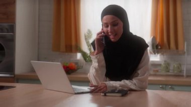 Mutlu gülümseyen Müslüman kadın tesettürlü Hintli bayan ev hanımı müşterisi dizüstü bilgisayarla yemek siparişi veriyor cep telefonuyla konuşuyor evde iletişim kuruyor.