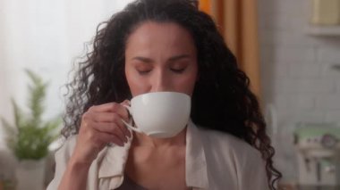 İspanyol beyaz kadın mutfak mutfağından zevk alıyor aromalı çay içiyor bitkisel kahve içiyor cappuccino latte espresso içiyor elinde kahve fincanı tutuyor ve sabah kahvaltısında kameraya gülümsüyor.