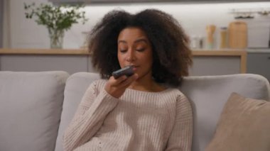 Mutlu Afro-Amerikalı kadın kanepeye oturmuş gülümseyen kadın cep telefonu hoparlörleri kullanarak sesli mesaj gönderiyor.