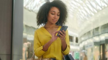 Genç Afrikalı Amerikalı kadın cep telefonuyla mesajlaşıyor alışveriş merkeziyle buluşuyor. Kara Cuma alıcı müşterisi modaya uygun bayan zımbırtıları, internet mesajları, akıllı telefon kullanıcıları, alışveriş kolikleri...