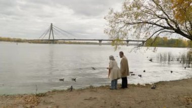 Arkaya bakan yaşlı kadın göl kenarında ördek besleyen ördek parçaları fırlatan yaşlı adam orta yaşlı yetişkin aile emekli eşler, erkek, kadın, sonbahar parkı şehrinde romantik bir randevu.