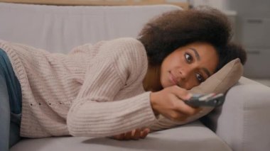 Afro-Amerikalı etnik kadın sıkılmış bir kız kanepede uzanıyor tembel kadın ev sahibi televizyon izliyor uzaktan kumandalı televizyon filmi izliyor evde gülümsüyor.