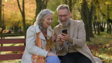 Yaşlı çift Kafkasyalı olgun kadın aile parktaki bankta cep telefonu kullanıyor neşeli eşler haberleri okuyor kahkahalar atıyor akıllı telefon kadın yaşlılık teknolojisi kullanıyor.