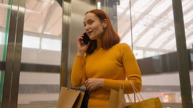 Mutlu, gülen, Kafkasyalı, kızıl, kadın, müşteri, modern cam asansöre binen tüketici, alışveriş çantaları, cep telefonları, telefon, ev içi alışveriş merkezleri.