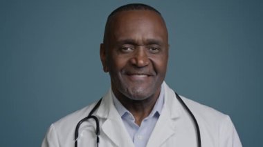 Yakın plan gülümseyen Afro-Amerikan erkek doktor stüdyo mavi arka plan etnik erkek cerrah doktor dişçi oftalmolog tıbbi hastane çalışanı kamera uzmanı