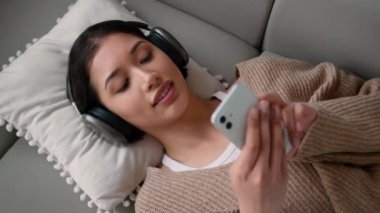 Kaygısız kadın ispanyol kız ev sahibi kanepeye uzanmış rahat rahat bir evde müzik kulaklıkları, cep telefonu ses uygulamaları rahatlayın hafta sonu kulaklık sesleri dinlendirici şarkılar