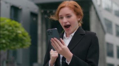 Happy şoke oldu, heyecanlı Kafkasyalı kızıl kadın iş kadını resmi takım elbise zaferi kutladı. Cep telefonu iyi haber. Mesajı kazan, iş başarısını şehir dışında kutla.