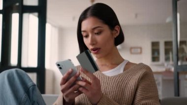 Kafkasyalı İspanyol tebessümlü kadın kredi kartı ve akıllı telefon sahibi mutlu bayan müşteri alıcısı indirimli cep telefonu ödemesi evde banka uygulaması hizmeti ile internet üzerinden alışveriş yapıyor.