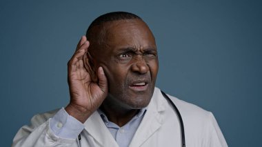Yakın plan kafası karışmış afro-amerikan erkek doktor stüdyo mavi arka planda el ele tutuşmuş kulak kaybı sorunu öne doğru eğil ve sessizce reklamları dinlemek için tıbbi test reklamlarını dinle.