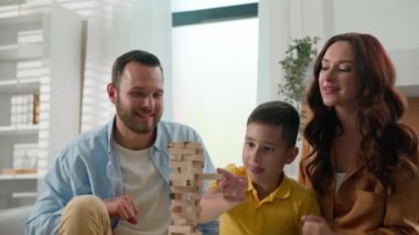 Neşeli, mutlu, beyaz aile dostu, şefkatli anne baba, oğluyla oyun oynuyor. Çocuk evde. Oturma odasındaki kanepede oturan ebeveynlerle, çocuklu tahta kule yapbozu oynuyorlar.