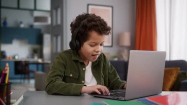 Küçük Afrikalı Amerikalı oğlan çoklu ırkçı çocuk öğrenci öğrenci öğrenci kulaklık takıyor komik video oyunu oynuyor sanal oyun oynuyor ev bilgisayarındaki bilgisayarı kullanarak zafer kazanıyor.