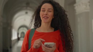 İspanyol mutlu öğrenci kız öğrenci kız koridorda yürüyor lisede üniversite koridorunda yürü sınıf eğitimine geç mutlu bir şekilde gülümse cep telefonu akıllı telefonu tut.