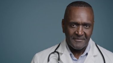Kendinden emin Afrikalı Amerikalı erkek doktor profesyonel sağlık çalışanı tıbbi temsilci cerrah kardiyolog onkolog mavi arka planda kamera görüntüsü veriyor.
