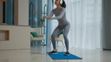 Beyaz tenli sporcu bayan sporcu sağlıklı zayıf atletik bayan gülen bayan evde egzersiz yapıyor sabah sporu yapıyor kulaklıkla müzik dinliyor vücuda çömeliyor