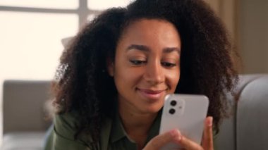 Kanepede akıllı telefon kullanan Afro-Amerikalı kadın gülümseyen bayan ev sahibi bağımlı cep telefonu koltukta uzanmış evde oturup mesajlaşarak internet üzerinden alışveriş cep telefonu uygulaması ile sohbet ediyor.