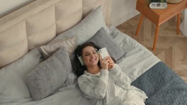 Genç, mutlu, esmer bir kadın, yatağında kulaklık takıp müzik dinliyor. Akıllı telefon teknolojisi kullanıyor. Keyifli, rahat, rahat, rahat bir kız.